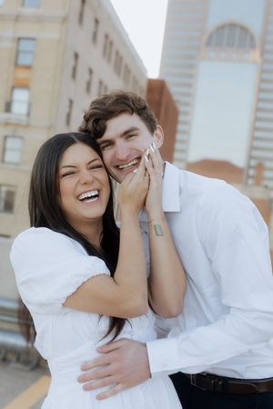 A couple wearing white shares a joyful moment laughing together in front of urban buildings.