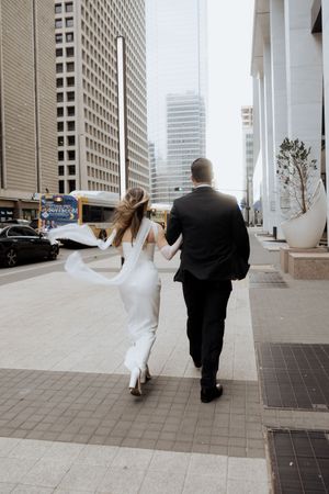 Couple in wedding attire walk hand in hand down a city sidewalk surrounded by tall buildings.