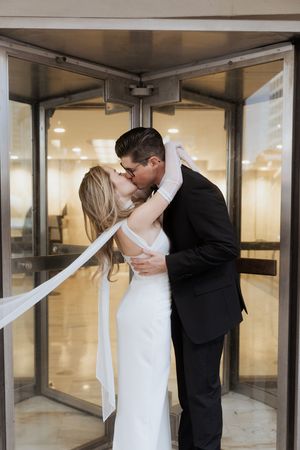 A romantic kiss shared between newlyweds in formal attire standing in a doorway with a white veil flowing.