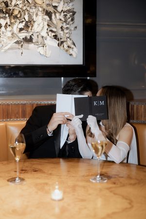 A couple on a dinner date holding up restaurant menus to hide behind while wine glasses sit on the wooden table.