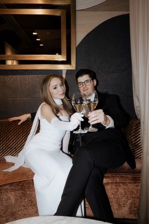 A newlywed couple in formal attire toasting with wine glasses while seated in an elegant restaurant booth.