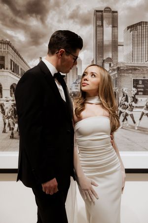 A romantic couple poses in formal attire against a cityscape backdrop in a sepia-toned photograph.