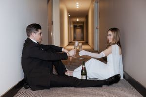 Two people sitting in a hotel hallway while sharing champagne and smiling on the carpeted floor.