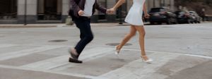 A couple in formal attire joyfully runs across a city crosswalk, the woman in a white mini dress and heels.