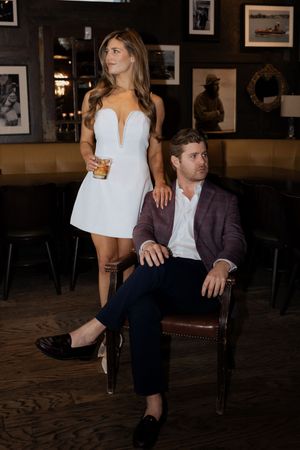 Elegant couple posing in a dark bar, woman in white mini dress holding a drink beside seated man in burgundy blazer.