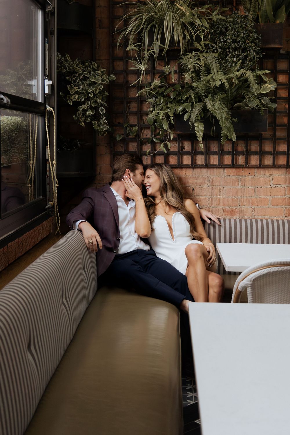 Couple sharing a romantic moment on a striped booth seat in a cozy restaurant with lush green wall plants.