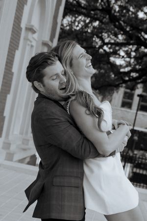 Black and white photo of a laughing couple embracing outdoors near a classic building, capturing a joyful romantic moment.