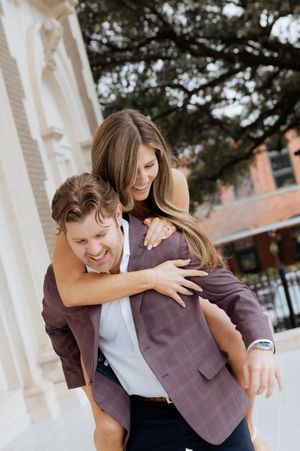 Happy couple laughing outdoors as woman hugs man giving her a piggyback ride near ornate columns.