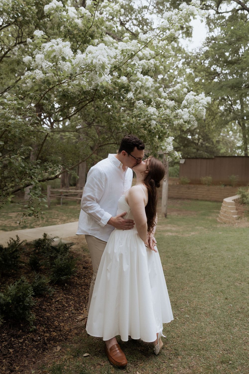 A couple shares a romantic moment under blooming white flowers in a garden setting, both dressed in elegant white attire.