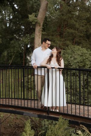 A romantic couple in coordinating white outfits stand intimately together on a wooden bridge in a lush forest setting.