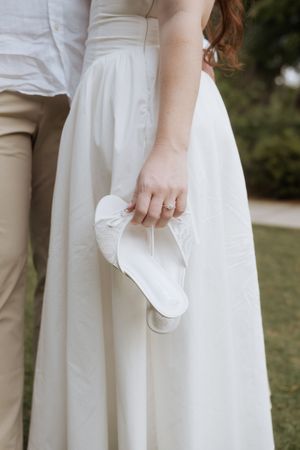 Hand holding white flat shoes against flowing white wedding dress and beige pants on grassy background.