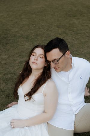 A tender moment between a couple dressed in white sitting together on grassy ground during a romantic photoshoot.