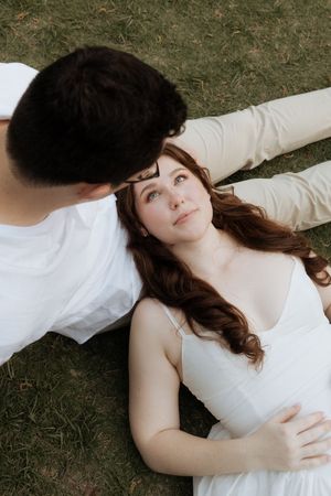 A couple in white outfits lies on grass together having an intimate moment while looking at each other.