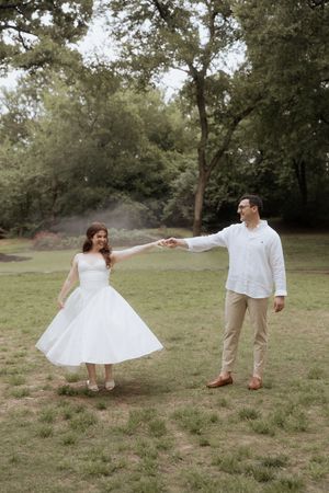 A couple in wedding attire stands in a grassy field with trees visible in the background, their legs and feet shown only.