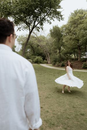 A woman in a white dress twirls on a grassy lawn as someone in white watches in a parklike setting.