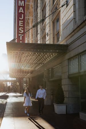 Two people walk beneath a vintage theater marquee with sunlight streaming through on an urban sidewalk.