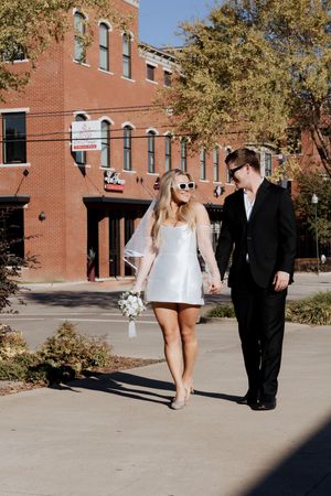 A couple in wedding attire walks together on a sidewalk in front of a historic brick building on a sunny autumn day.