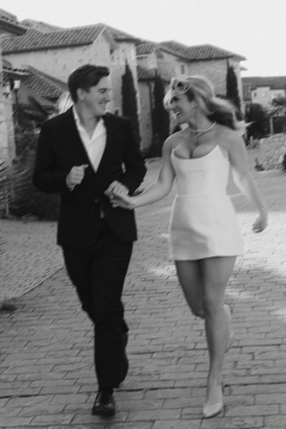 A playful staged conflict scene shows a woman in light clothing kicking at a man in a dark suit on a cobblestone street.