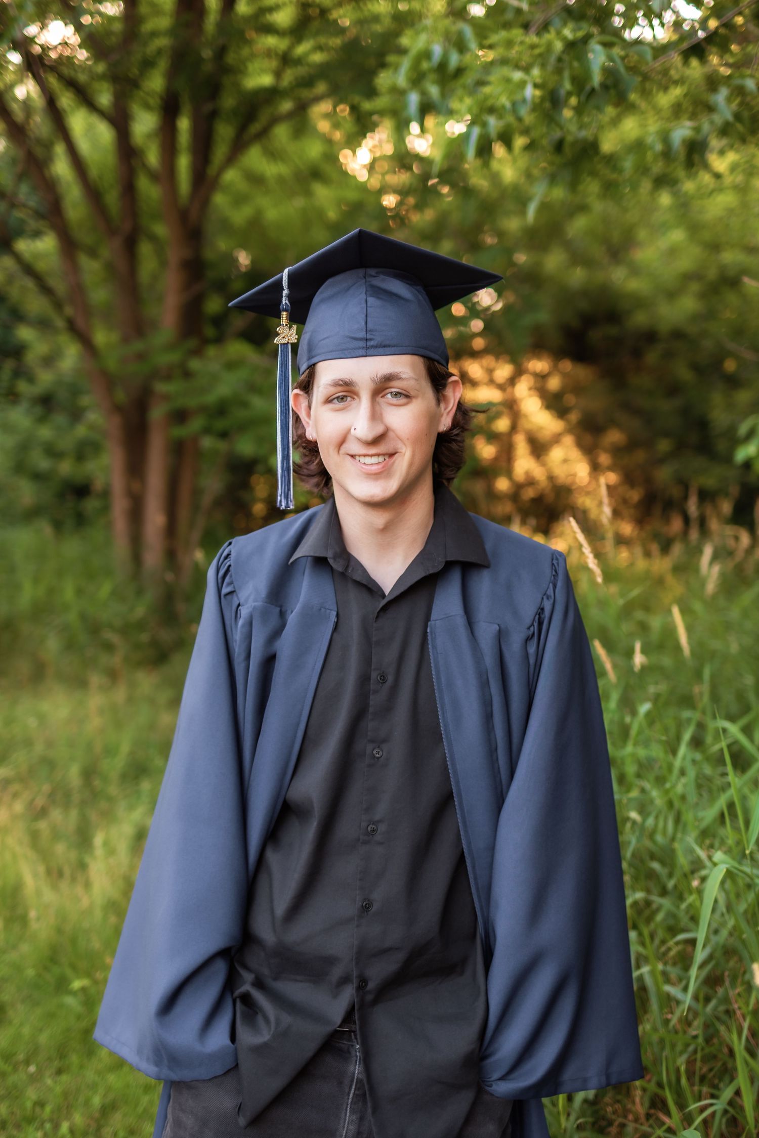 A graduate in traditional blue cap and gown poses outdoors against a green wooded background at sunset. Novi Michigan Senior Photographer
