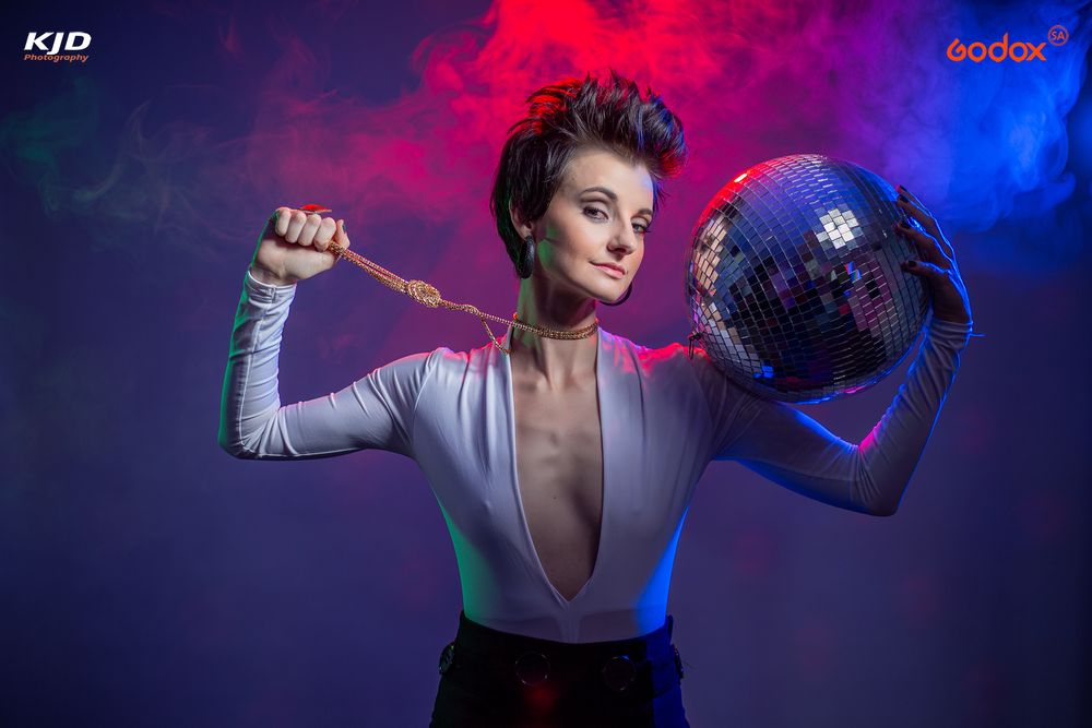 Figure holding a disco ball and glam rock sword pose against blue and pink smoky backdrop in dramatic lighting.