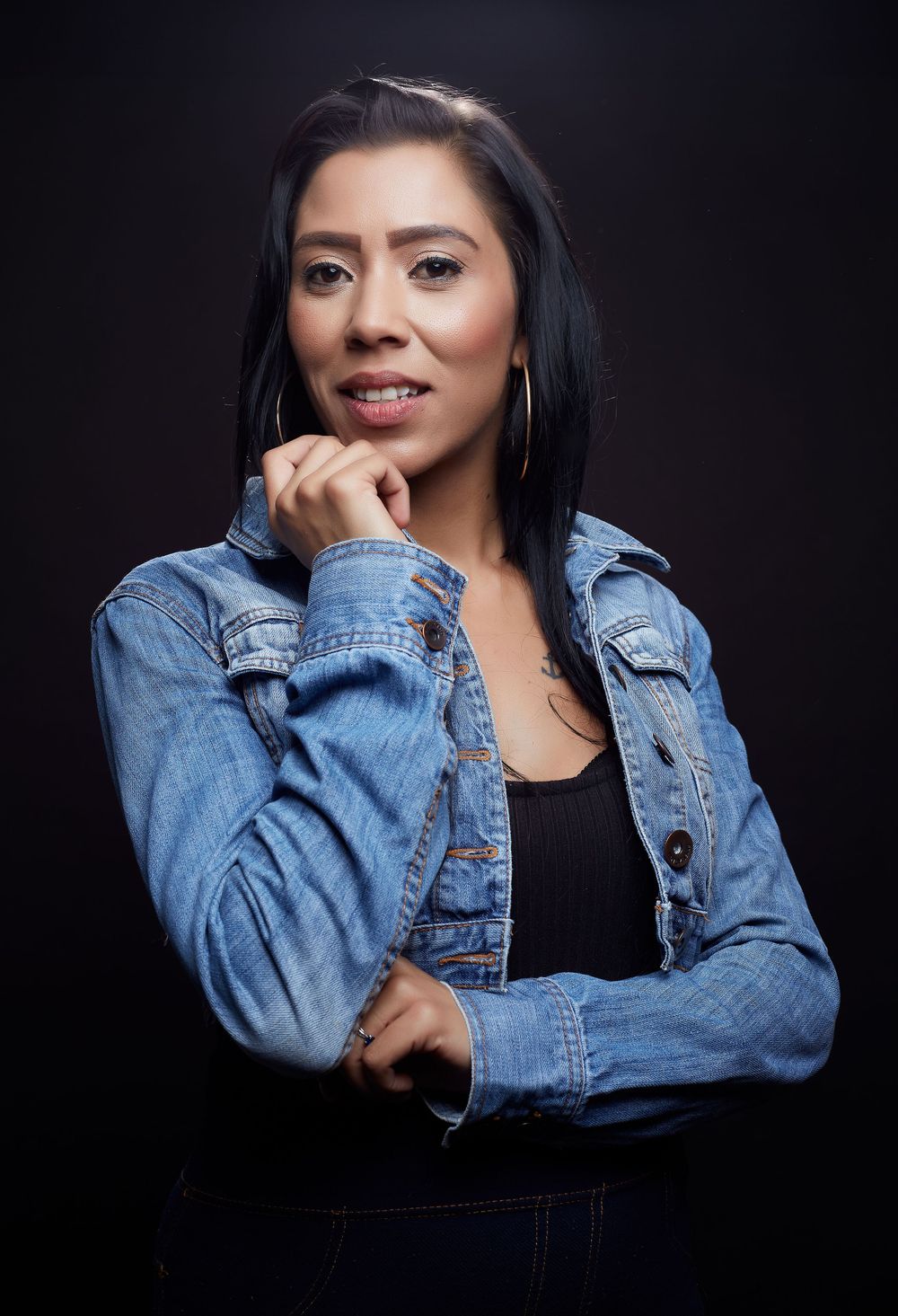 Portrait against dark backdrop showing confident pose in denim jacket and black top with silver hoop earrings.