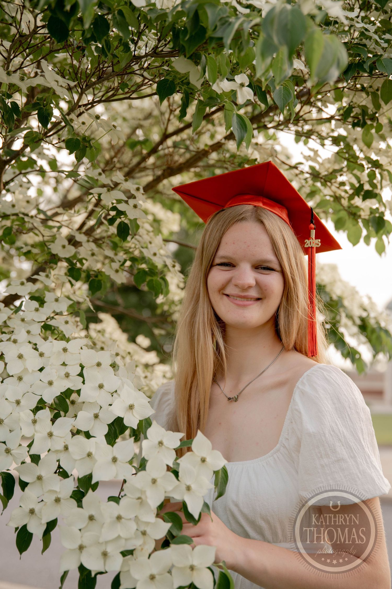 Graduate in red cap poses by blooming white dogwood flowers in white dress.