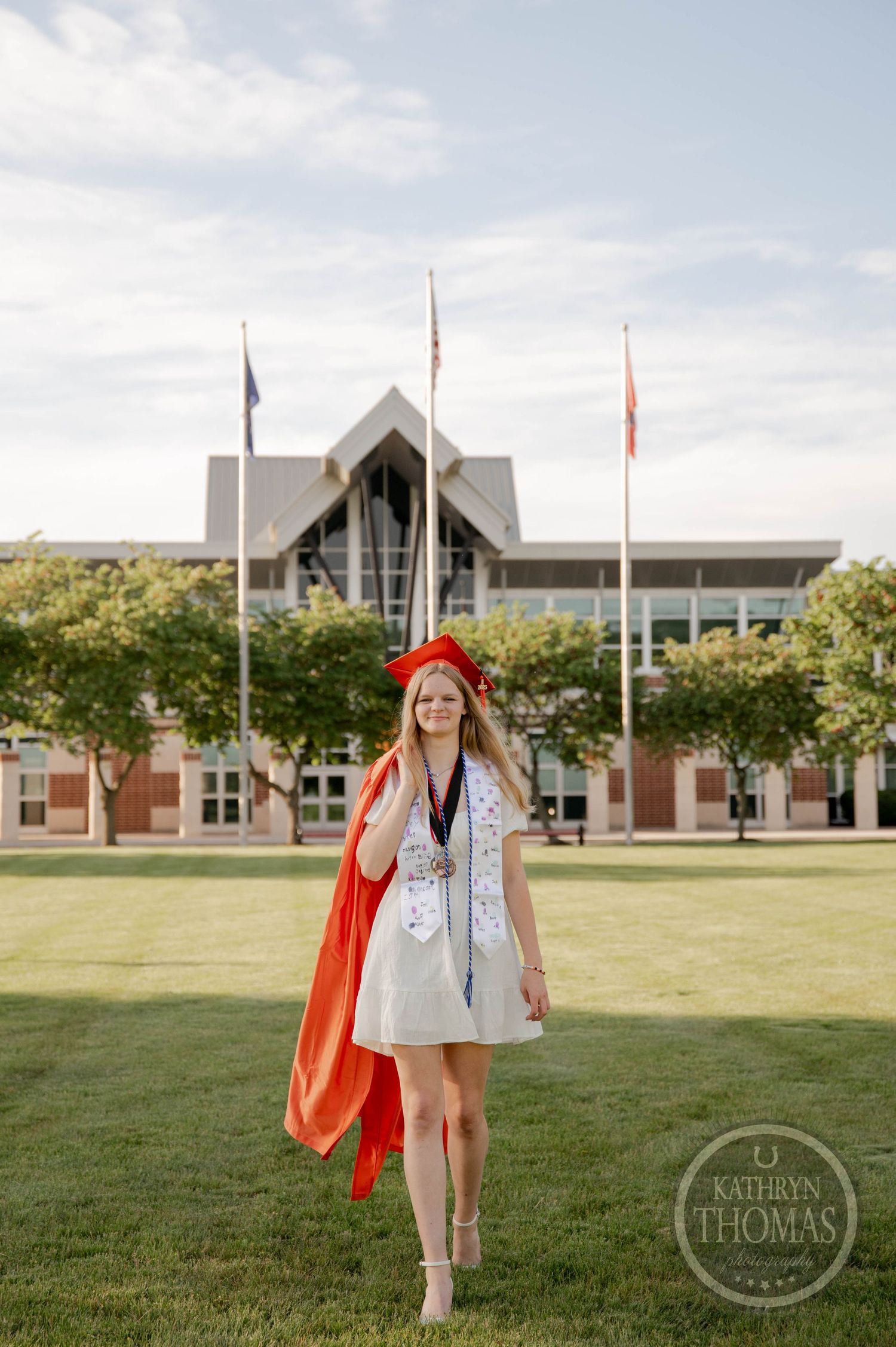 Graduate in orange and silver graduation attire poses on green lawn in front of academic building with flags.