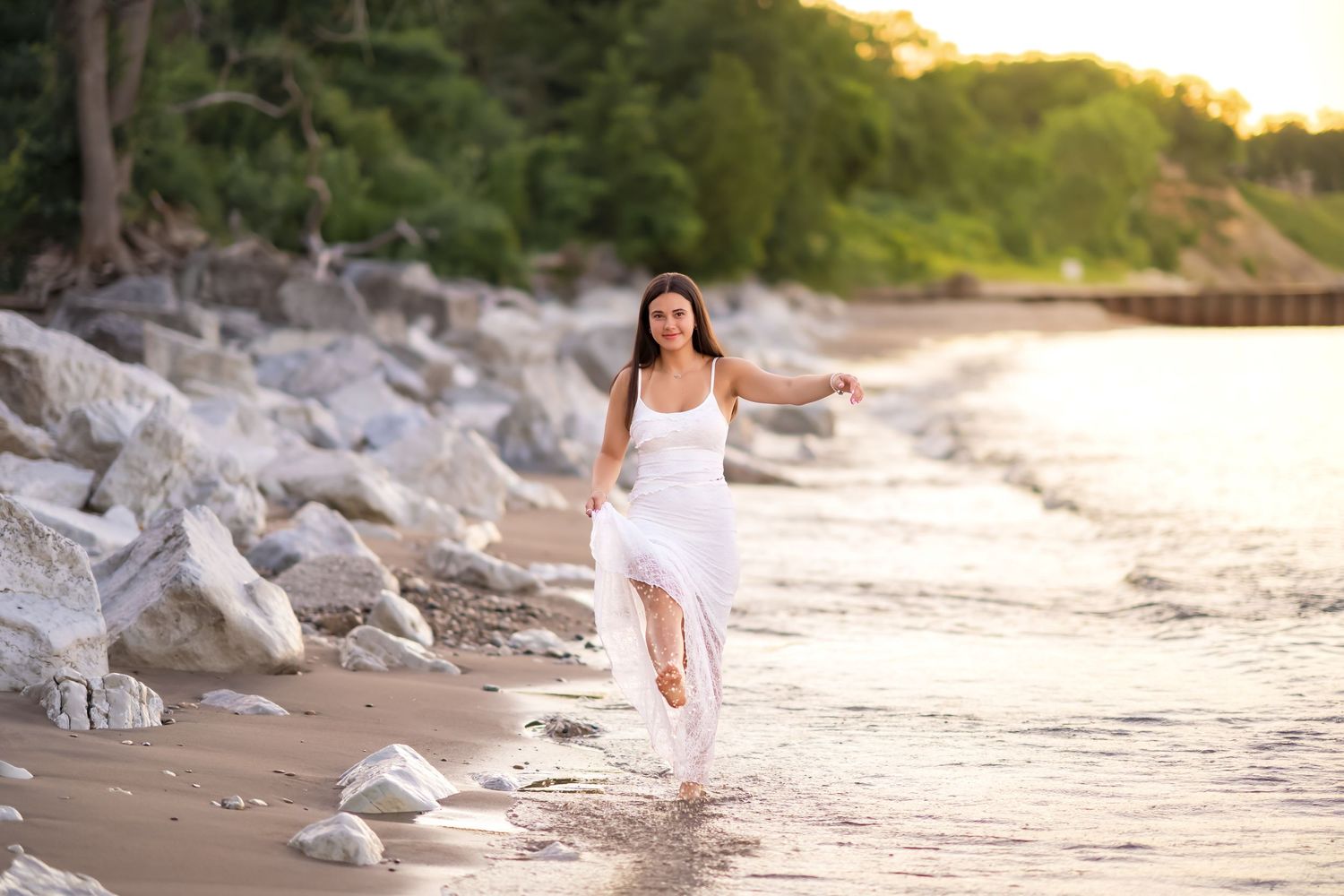 A person in a flowing white dress runs barefoot along a rocky beach shoreline at sunset.