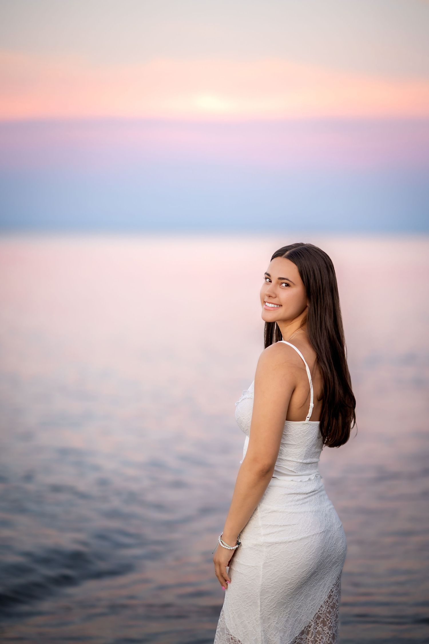 Soft pastel sunset colors over calm waters create a dreamy backdrop for a summer portrait session.