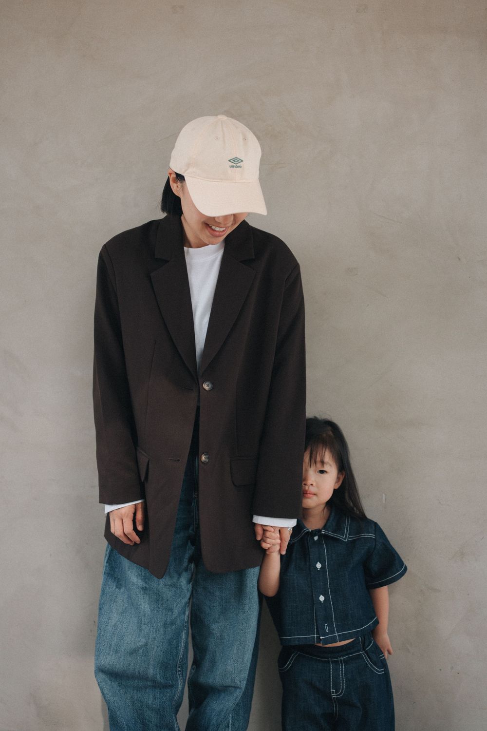 A parent and child pose together against a gray wall wearing denim and casual clothing with matching minimalist style.