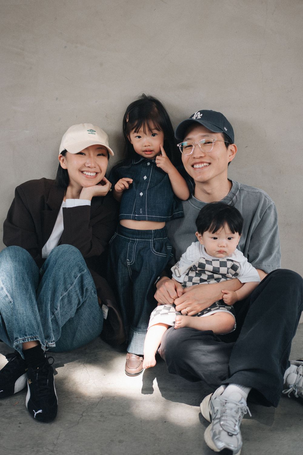 A casual family portrait against a grey wall shows four people sitting and smiling together.