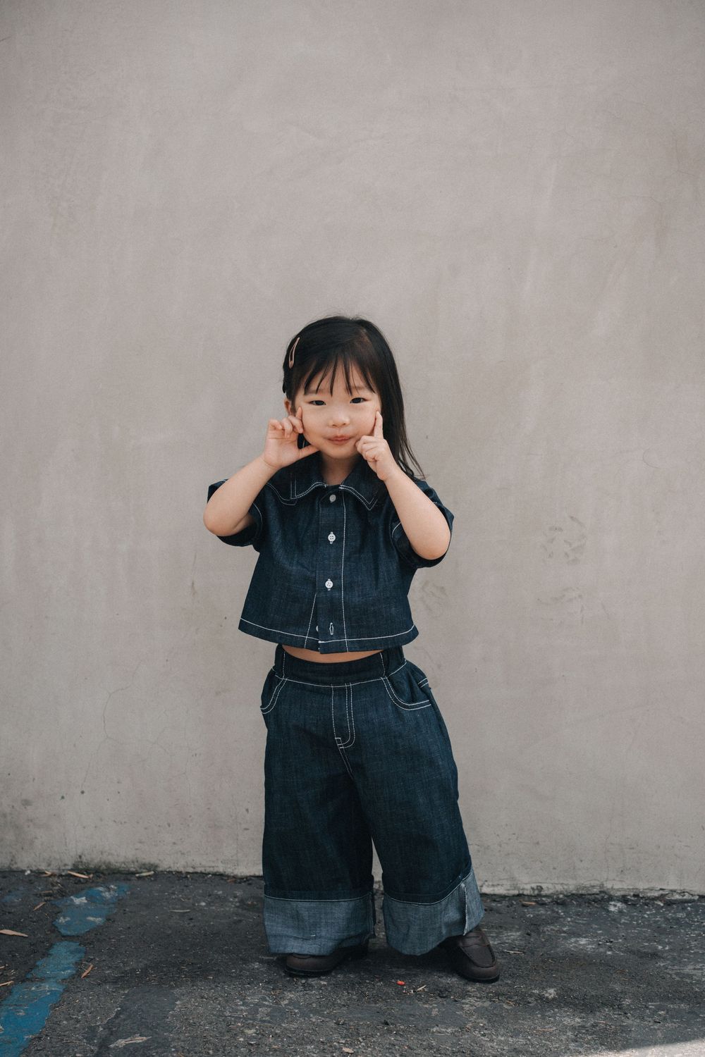 Child wearing matching denim outfit with crop top and wide-leg jeans poses playfully against gray wall.