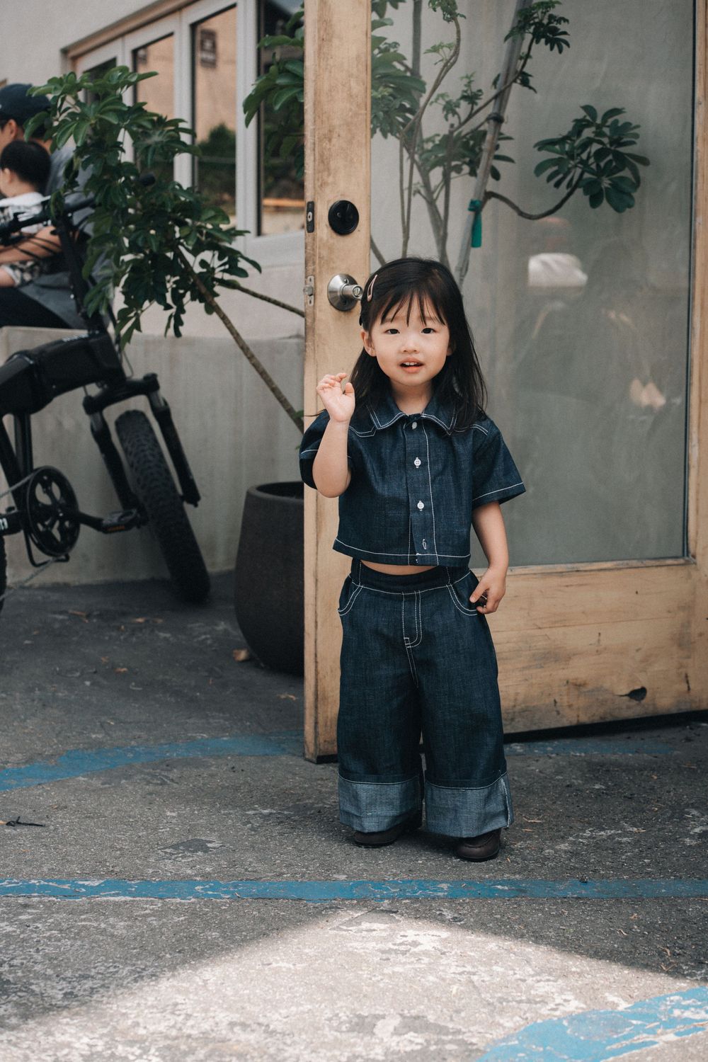 A toddler wearing a denim romper stands against a wall near a parked motorcycle in an urban setting.