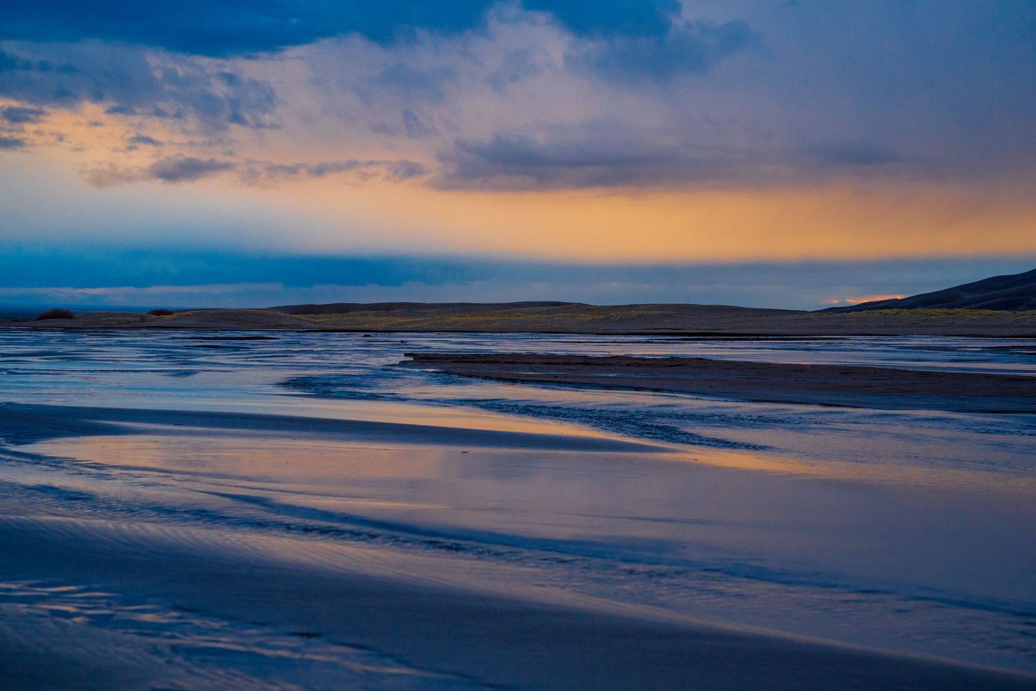 Dramatic sunset over a beach with reflective wet sand creating patterns along the shoreline. Great Sand Dunes National Park.