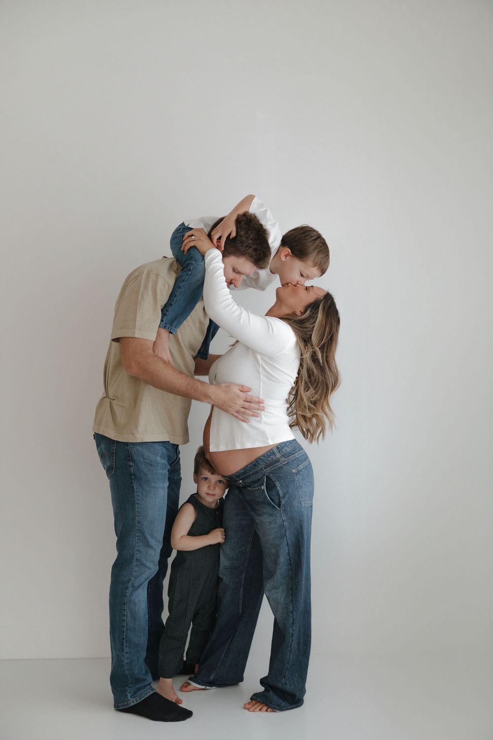 Pregnant woman kissing partner while holding child, with toddler hugging her legs, family maternity photo on white background.