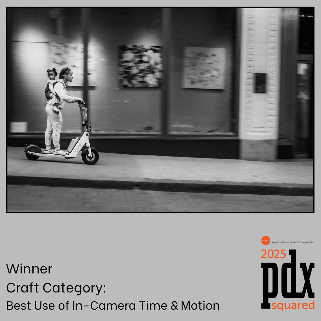 Black and white photo of someone riding an electric scooter past storefronts, creating motion blur effect on city street.