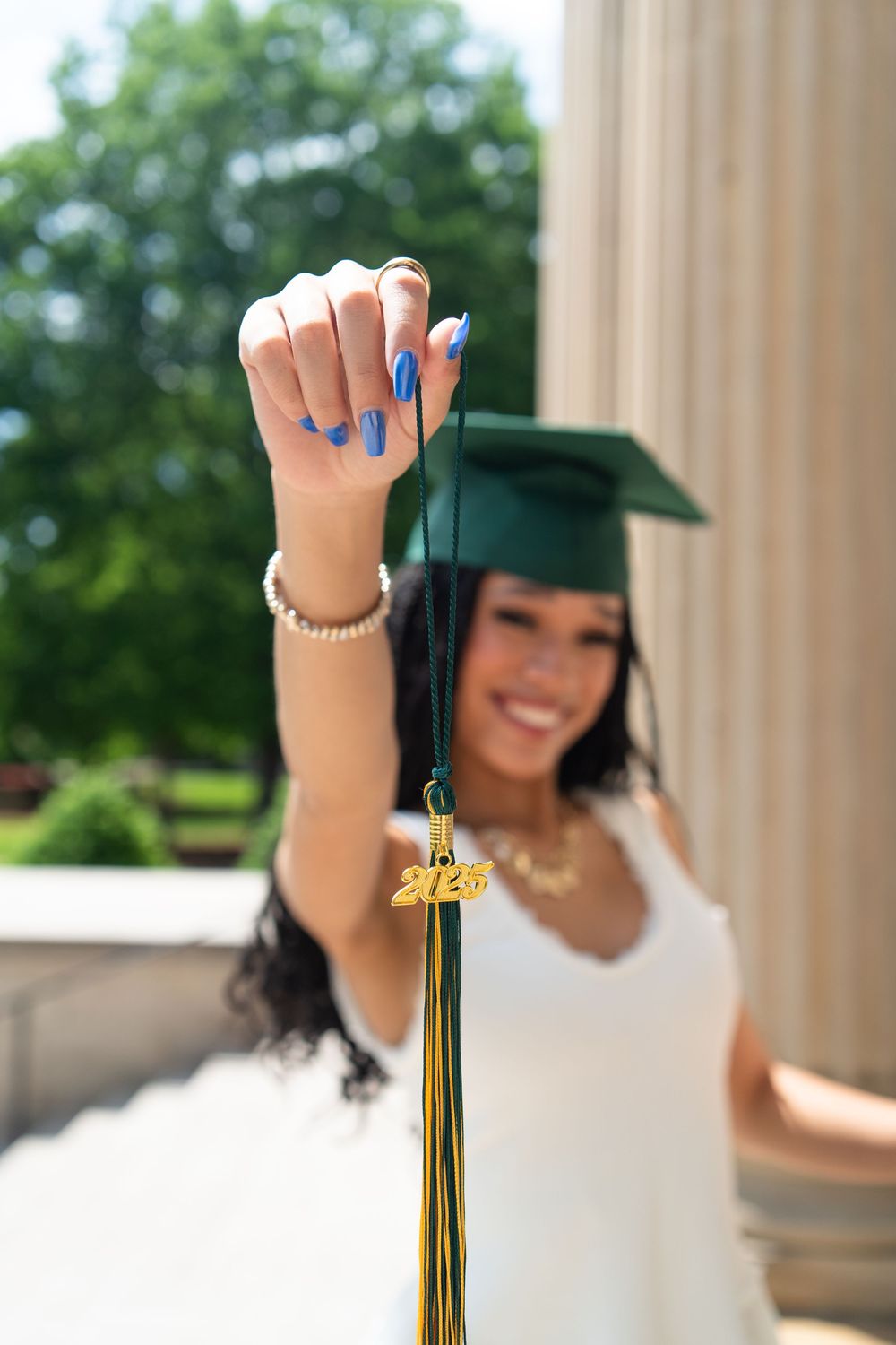 Graduate in white dress holds up tassel and graduation cap cord with painted blue nails against outdoor background.