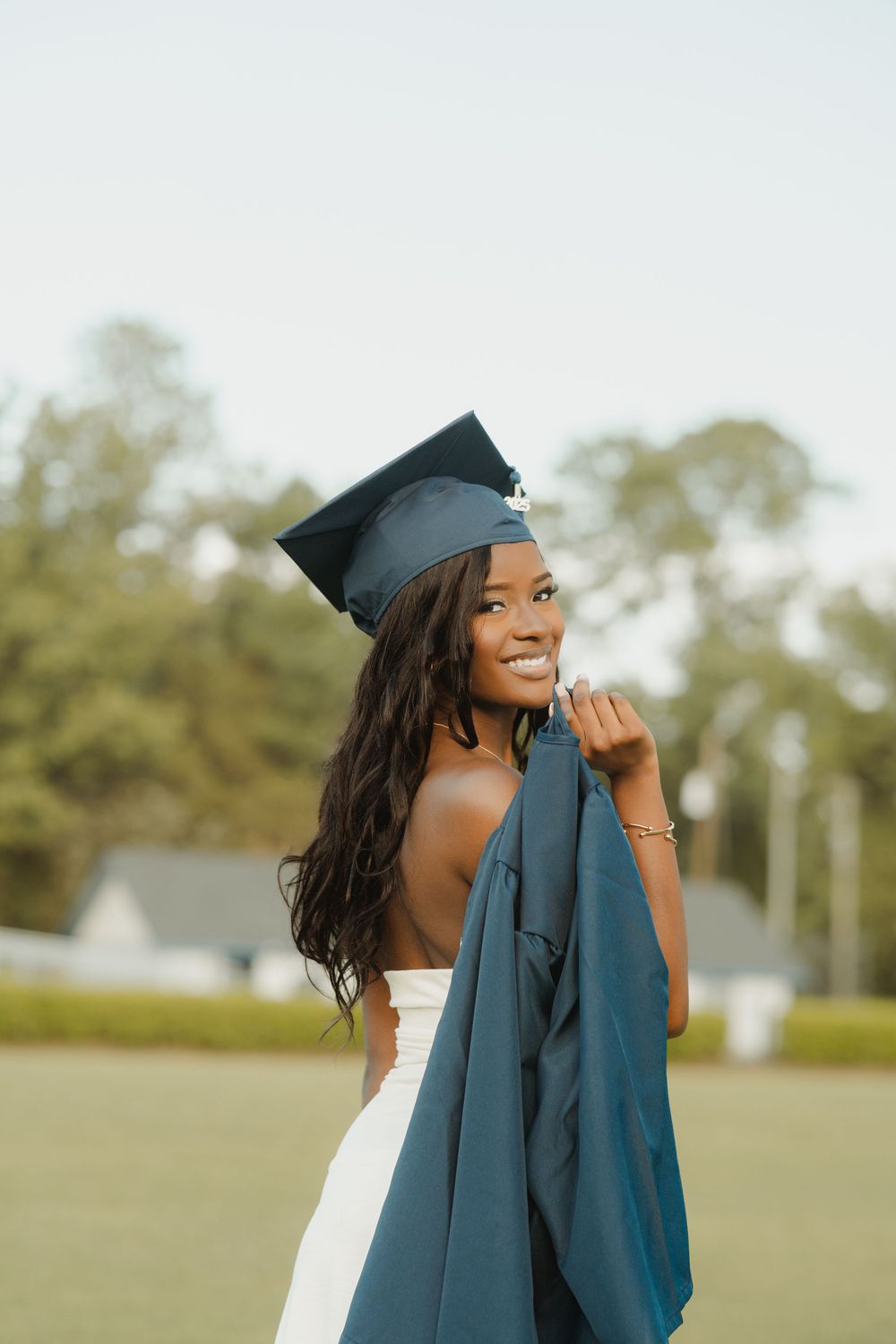 Graduating student wearing cap and white dress poses outdoors with academic gown against blurred natural background.