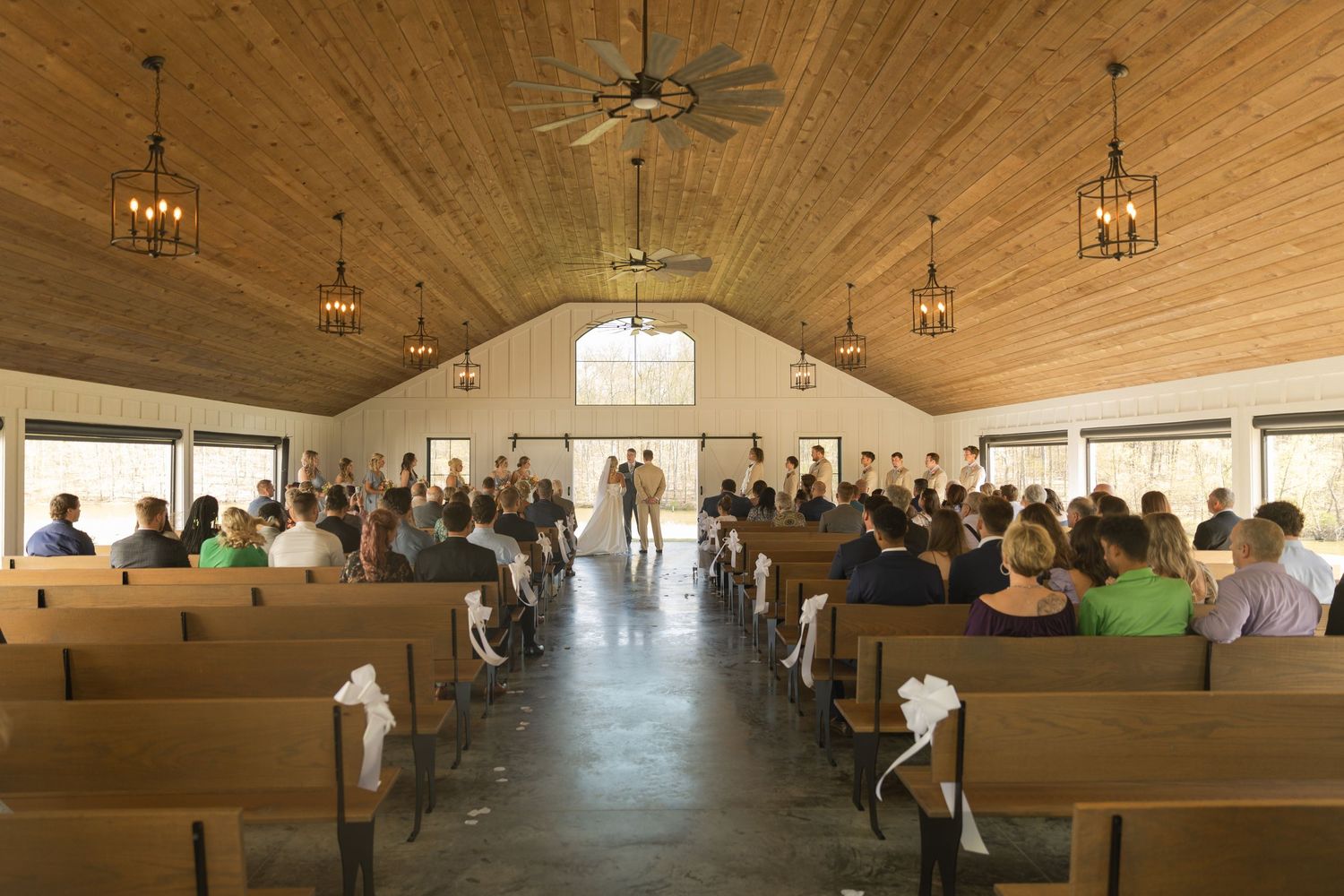 Interior of a rustic chapel with wooden pews and ceiling during a gathering, illuminated by hanging lanterns and natural light.