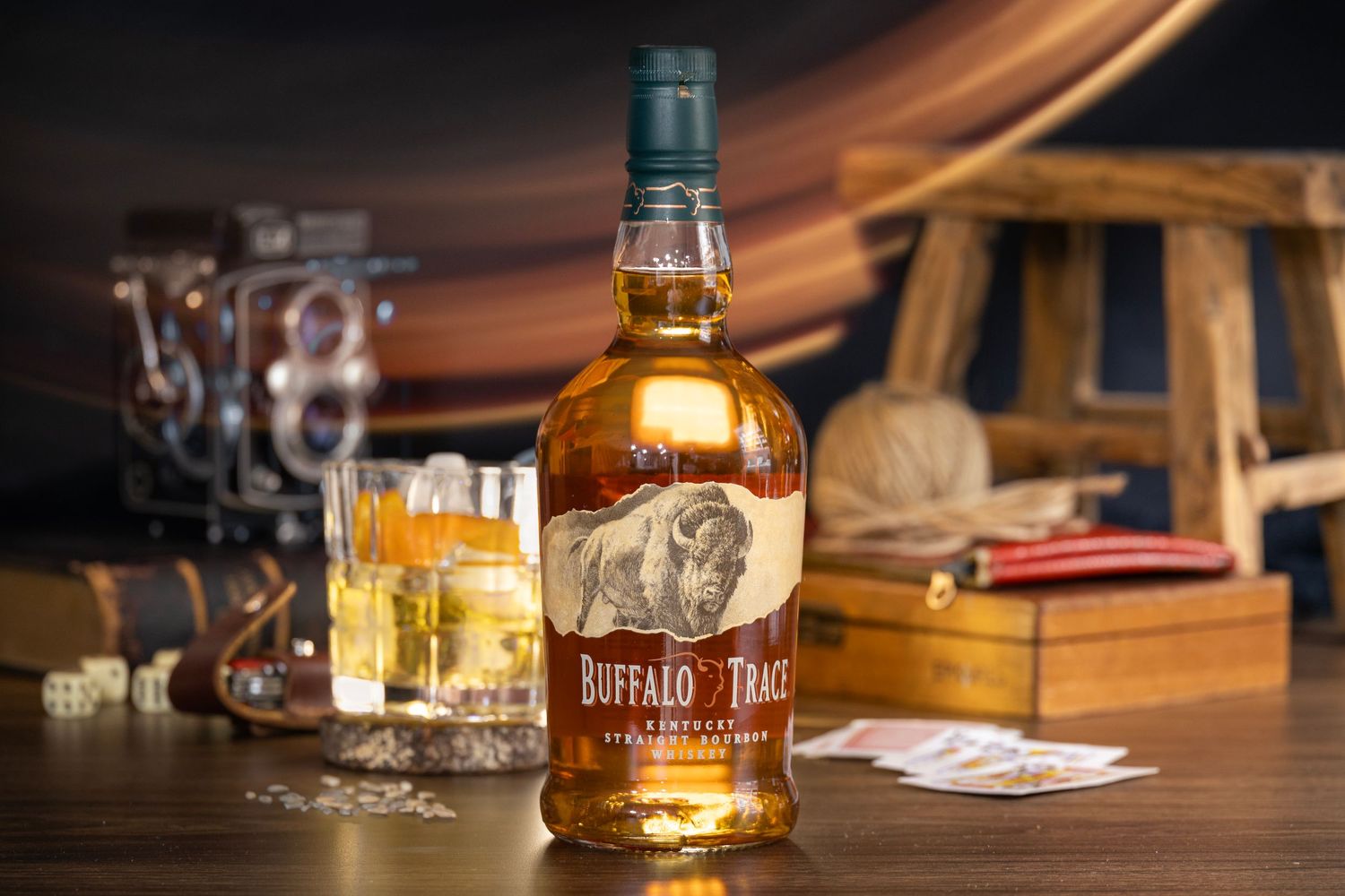 Buffalo Trace bourbon bottle beside a glass with ice, vintage camera, dice, cards, and wooden decor in a rustic bar setting.