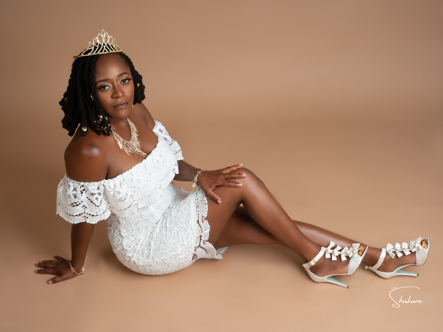 A formal portrait against a brown backdrop features someone in a white lace dress and tiara wearing white sneakers.
