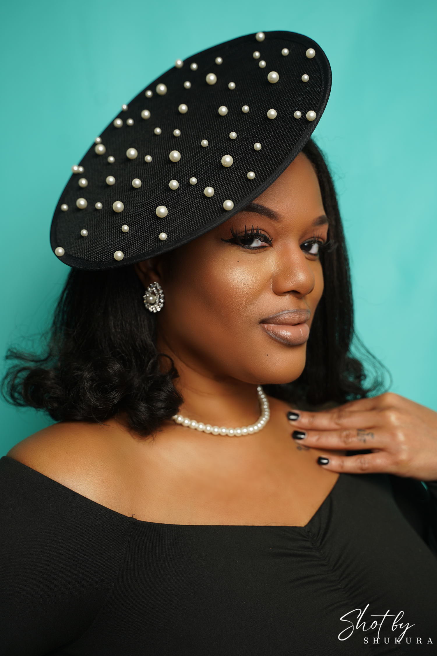 Pearl studded black beret styled with pearl necklace and black off-shoulder top against turquoise background.