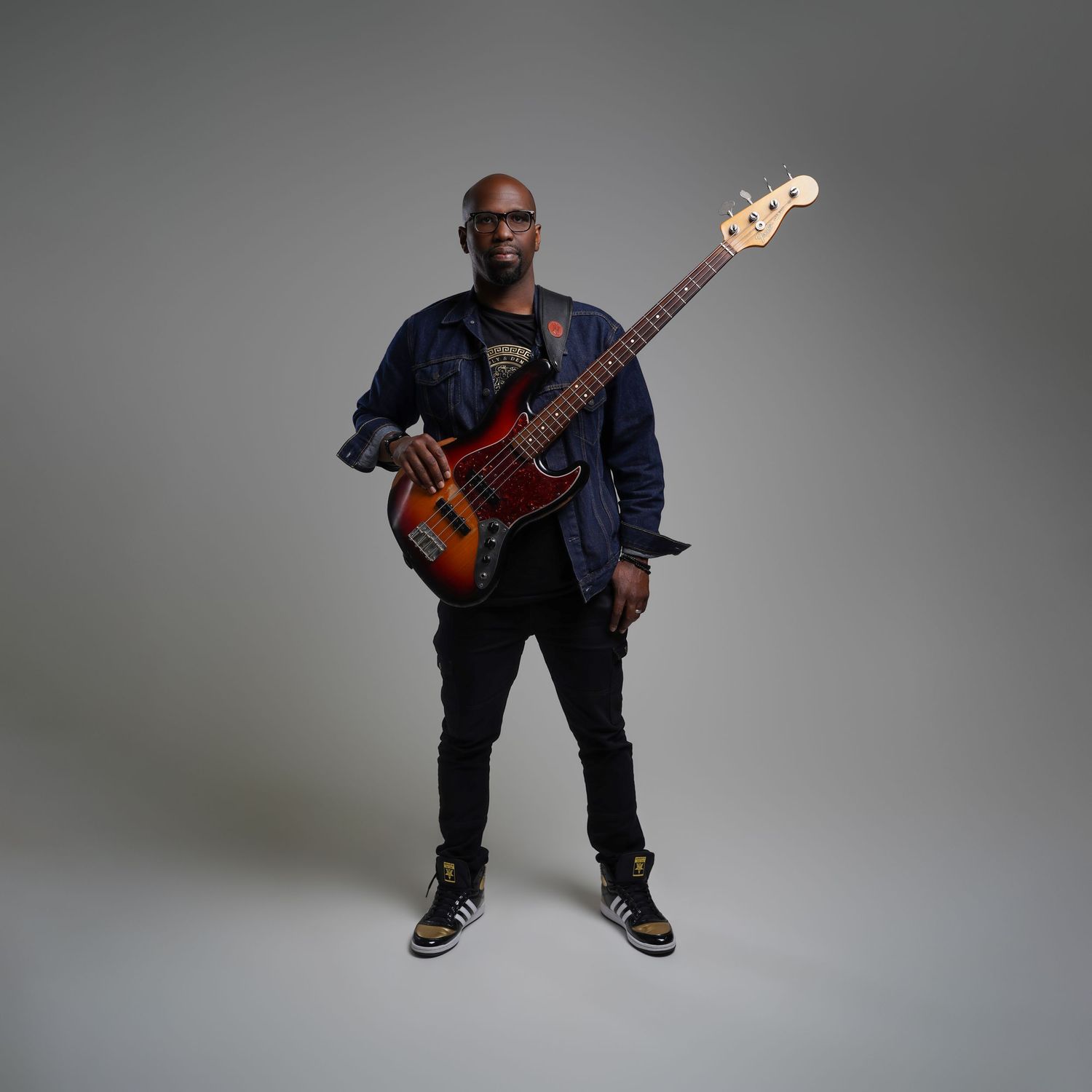 A musician in dark clothing holds a brown bass guitar while posing against a gray studio backdrop.