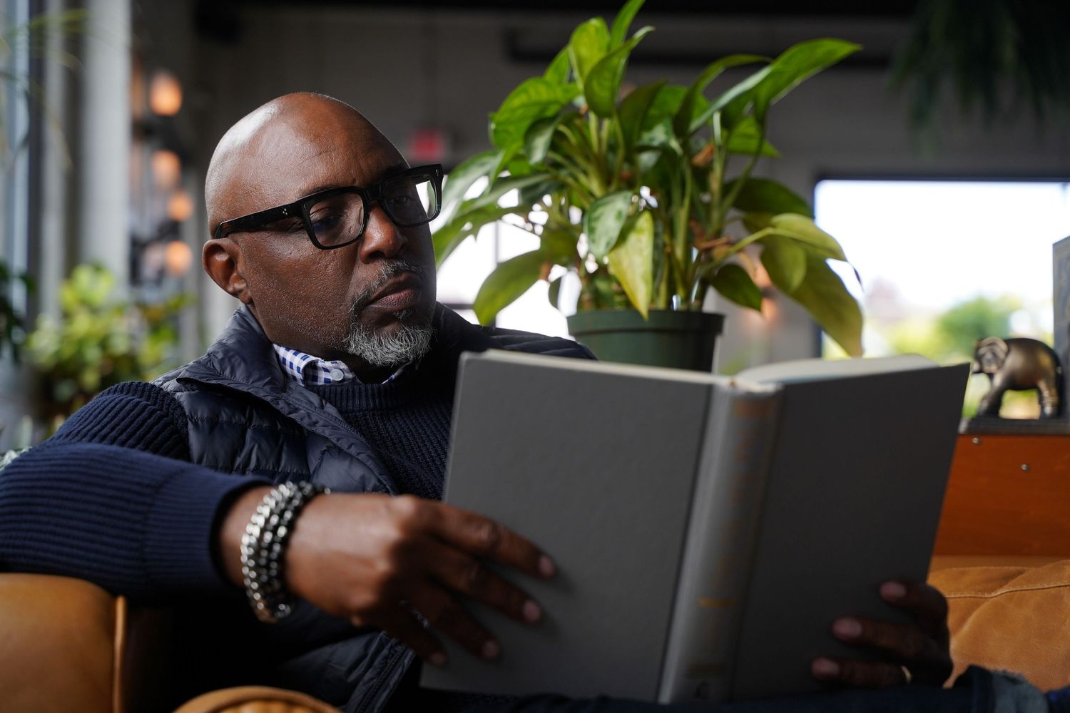 Someone wearing glasses and bracelet reads a book while seated by houseplants in a cozy indoor setting.