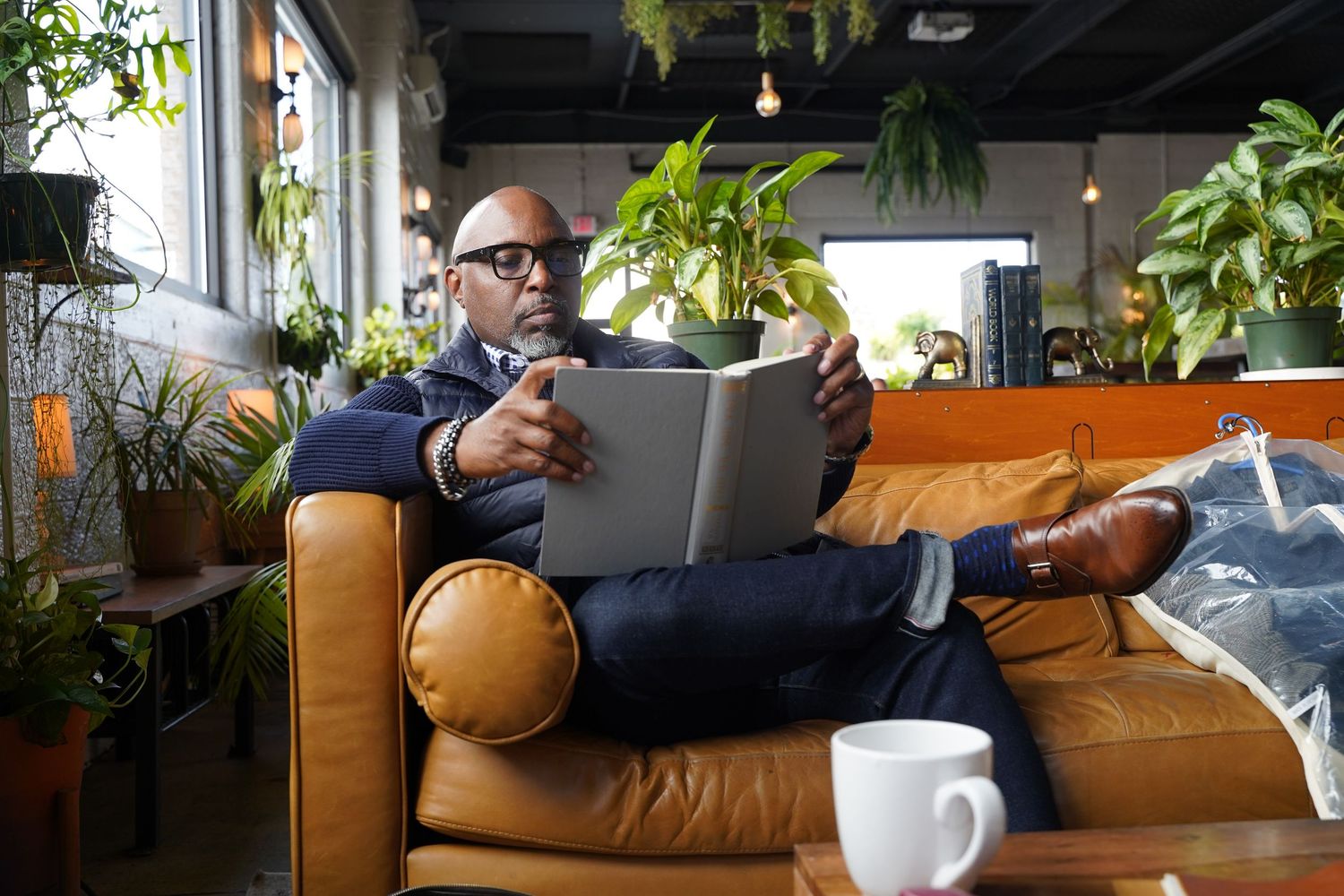 Someone relaxes and reads a book on a tan leather couch in a plant-filled room with a coffee cup nearby.