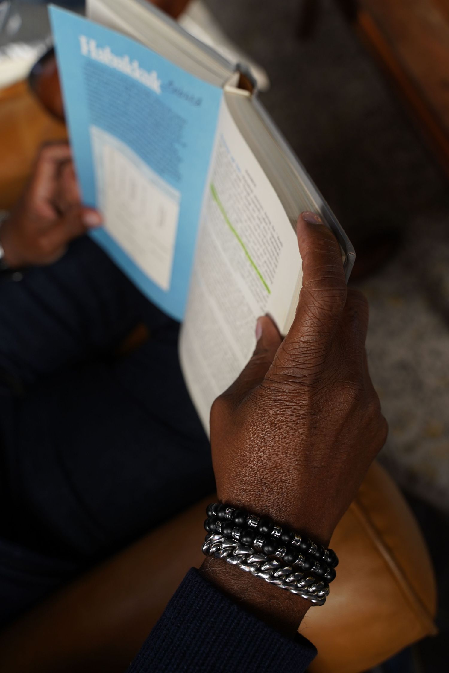 A hand wearing layered chain and beaded bracelets holds an open light blue book against a dark background.