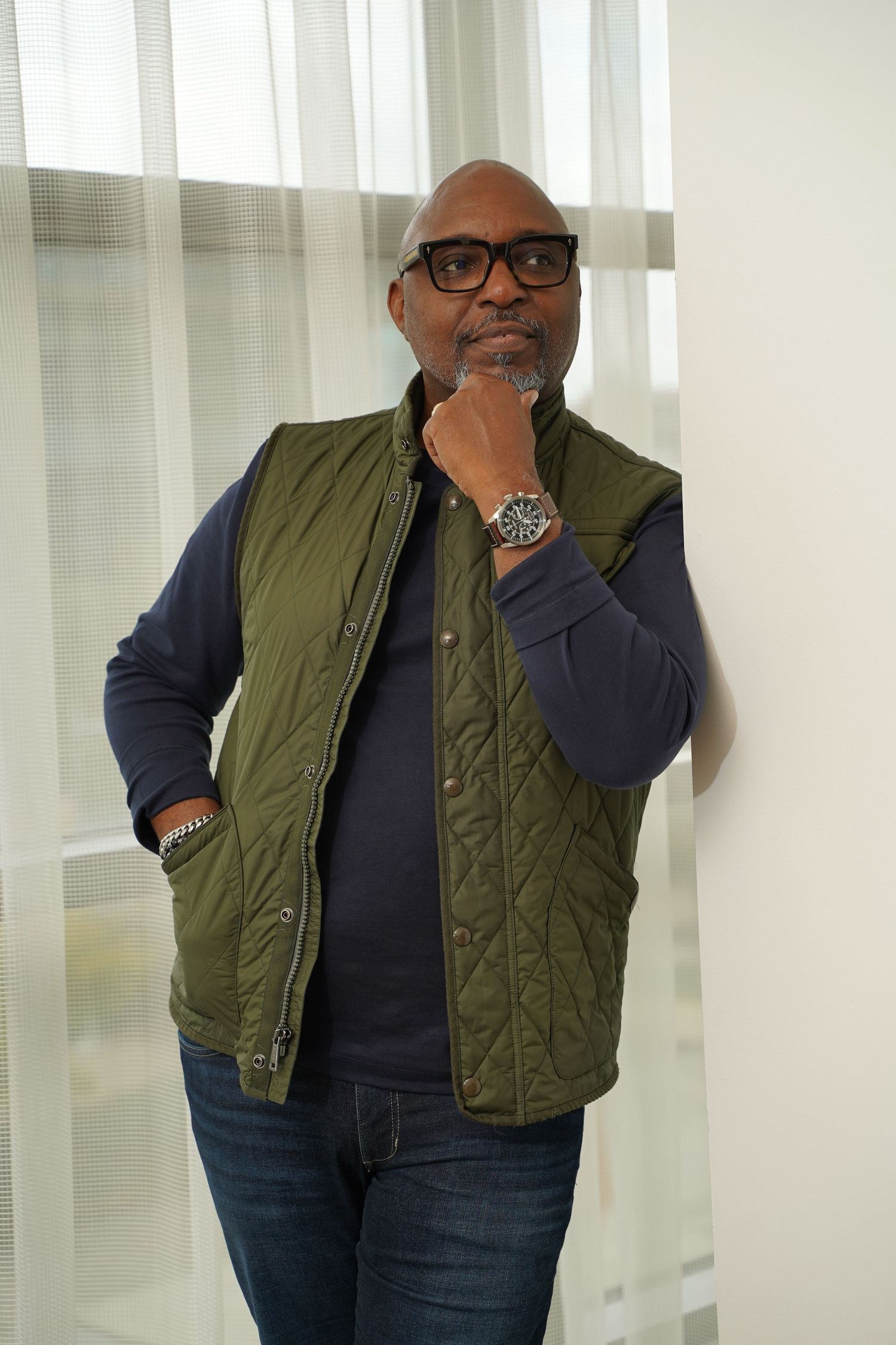 Man wearing black-rimmed glasses poses thoughtfully in an olive quilted vest and navy sweater by white curtains.