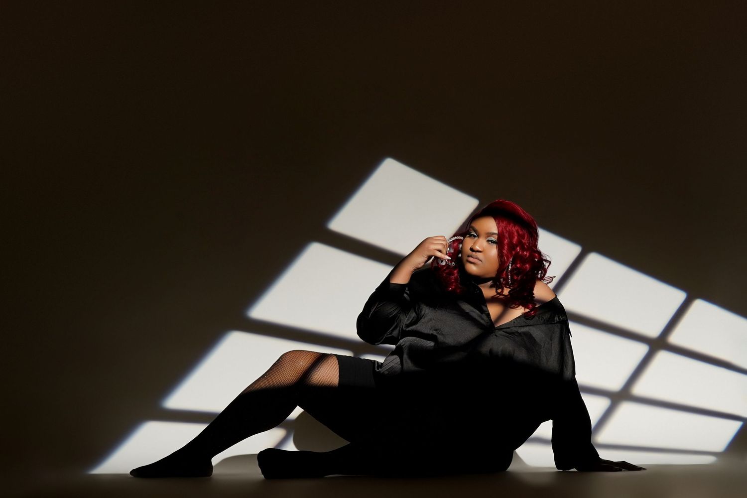 Person with red hair sits on floor in dramatic lighting with geometric window shadows cast on wall behind them.