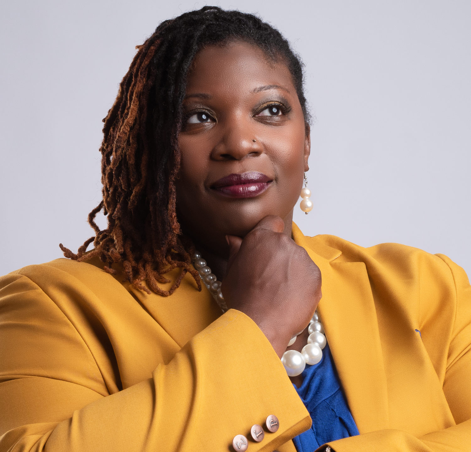 Professional portrait of someone in a mustard yellow blazer and blue top with pearls and gold earrings.