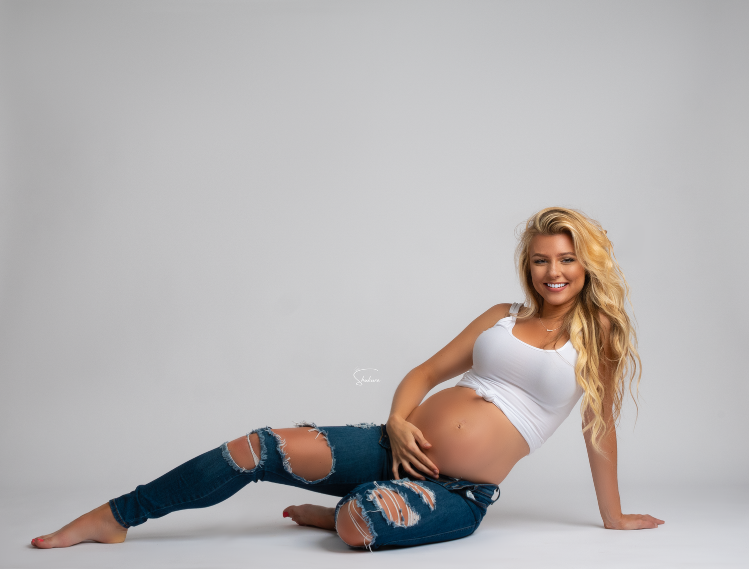 Expecting mother in white tank top and ripped jeans poses casually while sitting on floor against grey background.